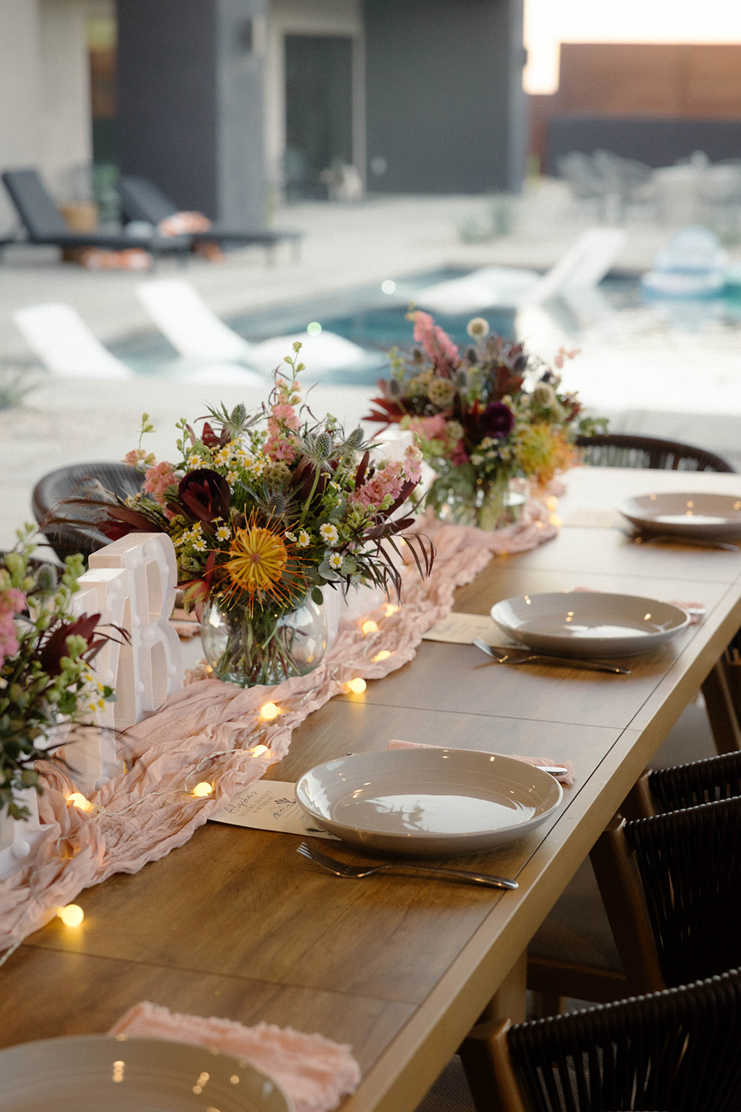 Long poolside table with blush runner, wildflower arrangements and string lights at sunset