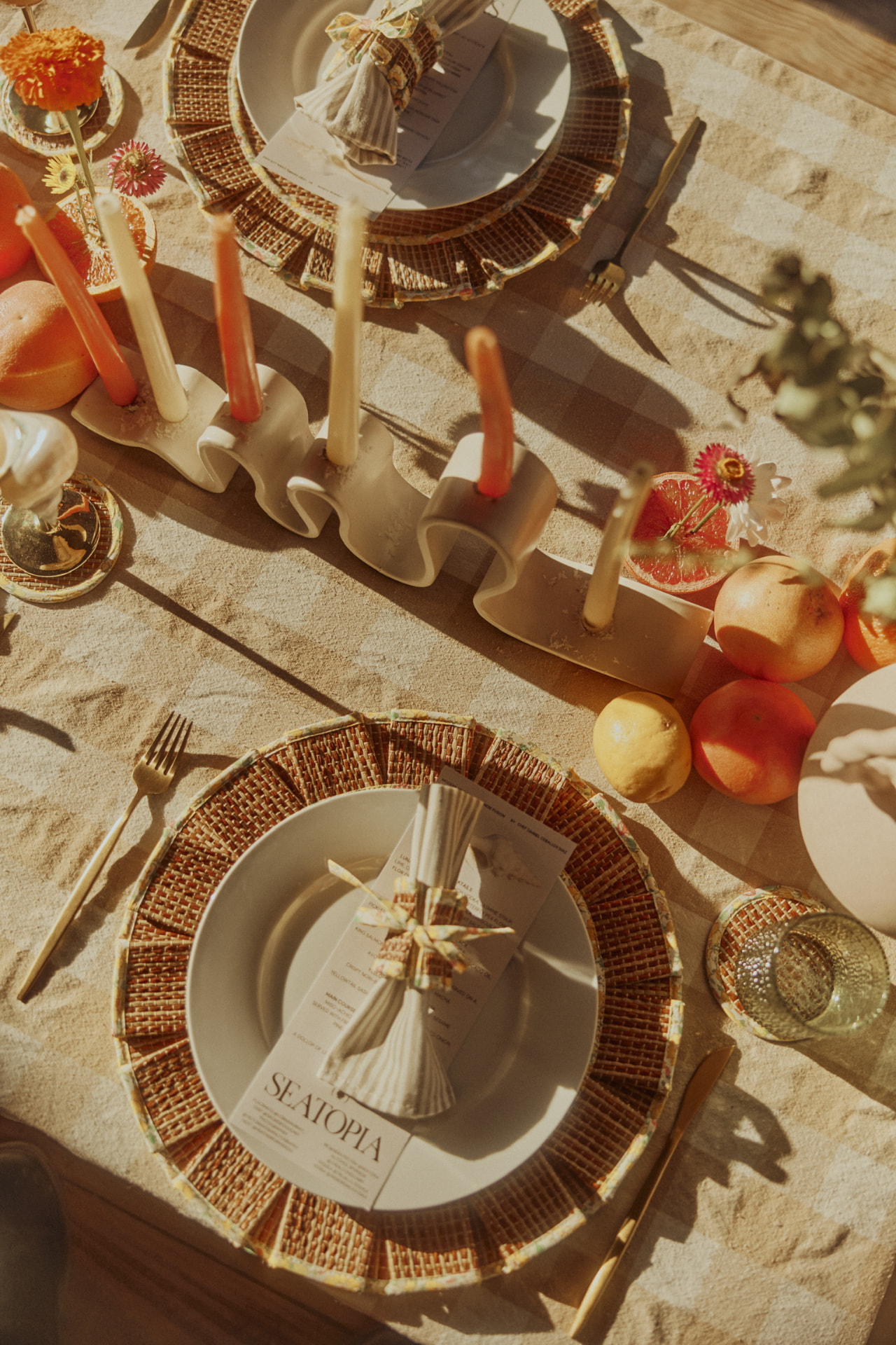 Overhead view of a checkered tablecloth with woven chargers, tapers and citrus
